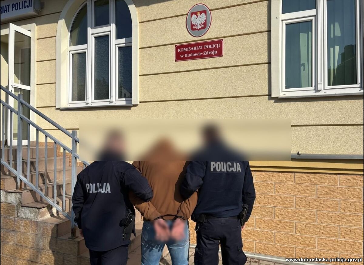Dwóch policjantów w mundurach prowadzi zakutego w kajdanki mężczyznę do budynku Komisariatu Policji w Kudowie-Zdroju