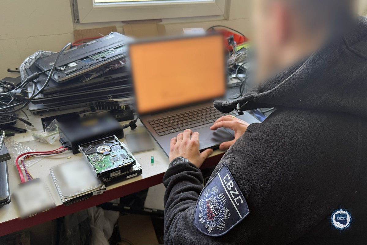 Funkcjonariusz CBZC w czarnej kurtce z naszywką pracuje przy laptopie podczas operacji Hellfire, na biurku rozłożony sprzęt elektroniczny