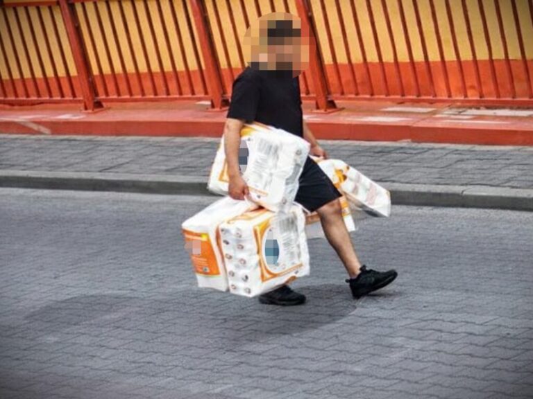 Mężczyzna z zamazaną twarzą niesie po chodniku kilka dużych, zgrzanych opakowań papieru toaletowego.