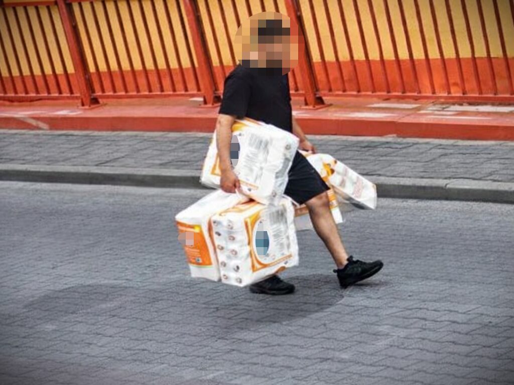 Mężczyzna z zamazaną twarzą niesie po chodniku kilka dużych, zgrzanych opakowań papieru toaletowego.